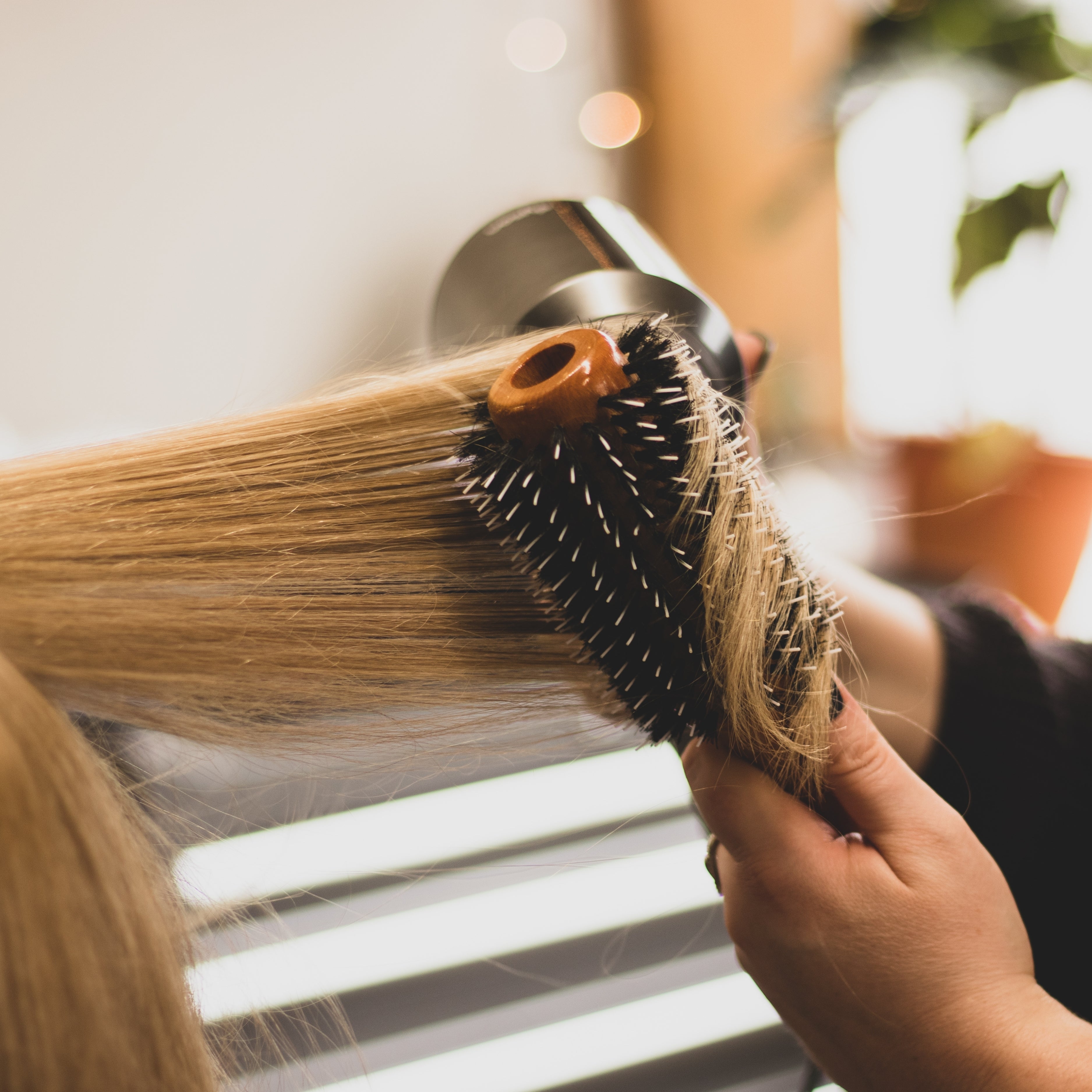 A close up image shows a hair stylist performing a blow-out on a woman's hair with a blow dryer and round bristle brush