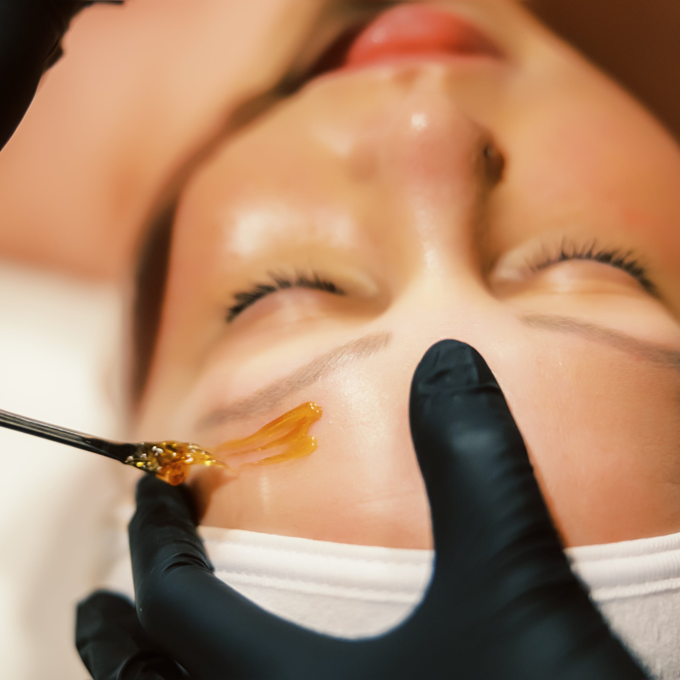 A close-up of a woman with eyes closed receiving an eyebrow sugaring treatment.
