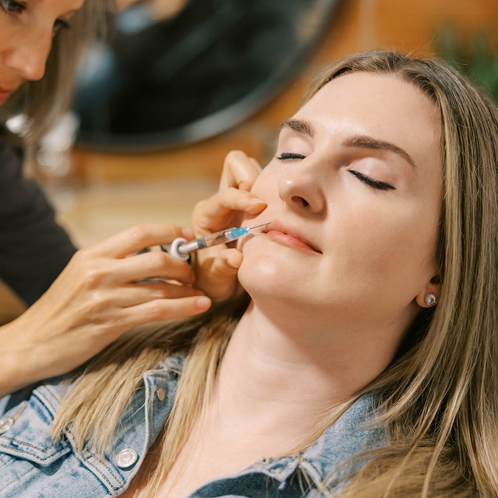 A woman relaxes with her eyes closed in a luxury spa setting while a registered nurse injector administers dermal filler.