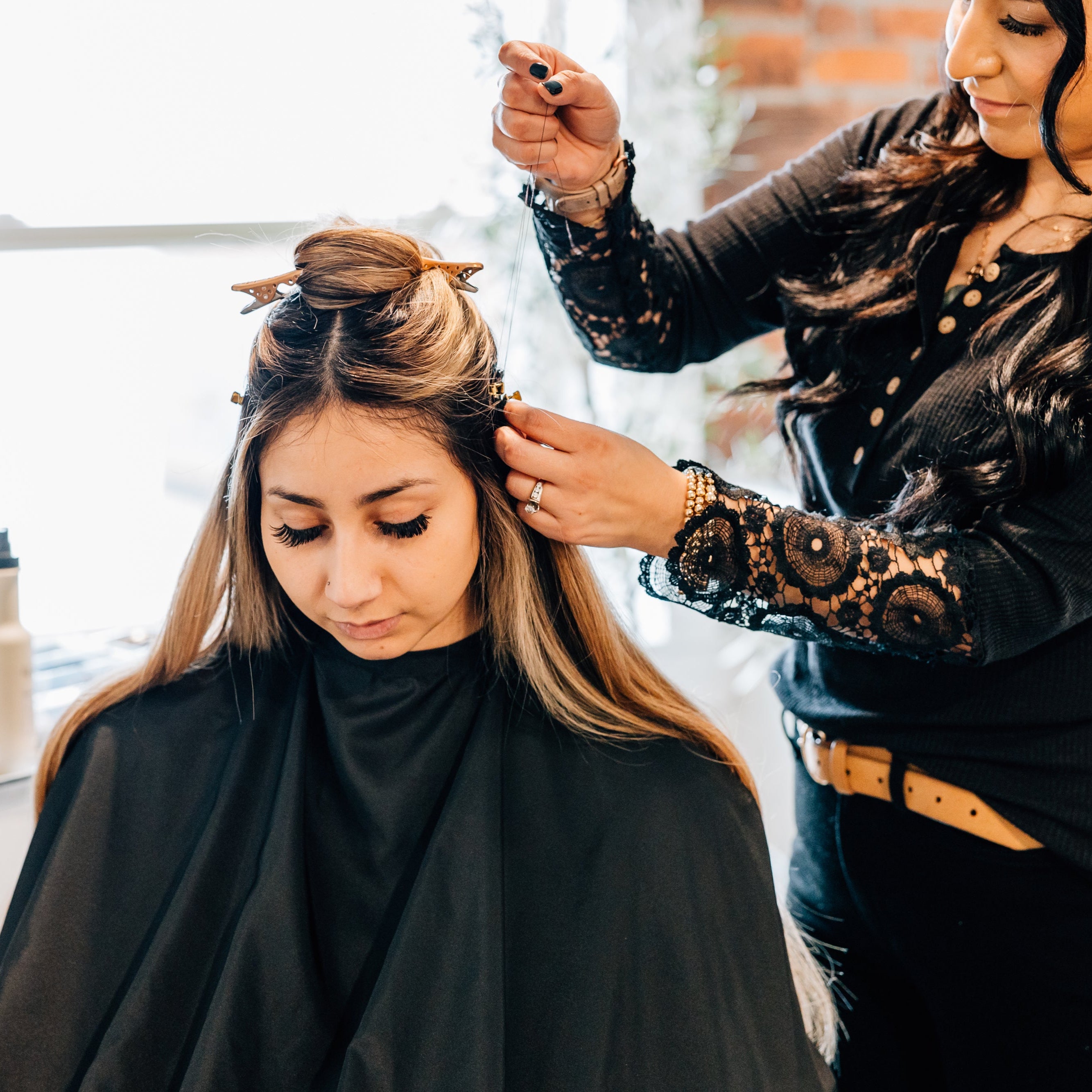 A hair stylist installs hand-tied hair extensions into a woman's hair in a luxury spa setting