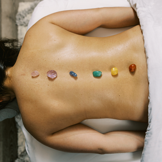 A woman relaxes on a massage table in a luxury spa setting while receiving reiki energy work with six elemental stones resting along her spine.