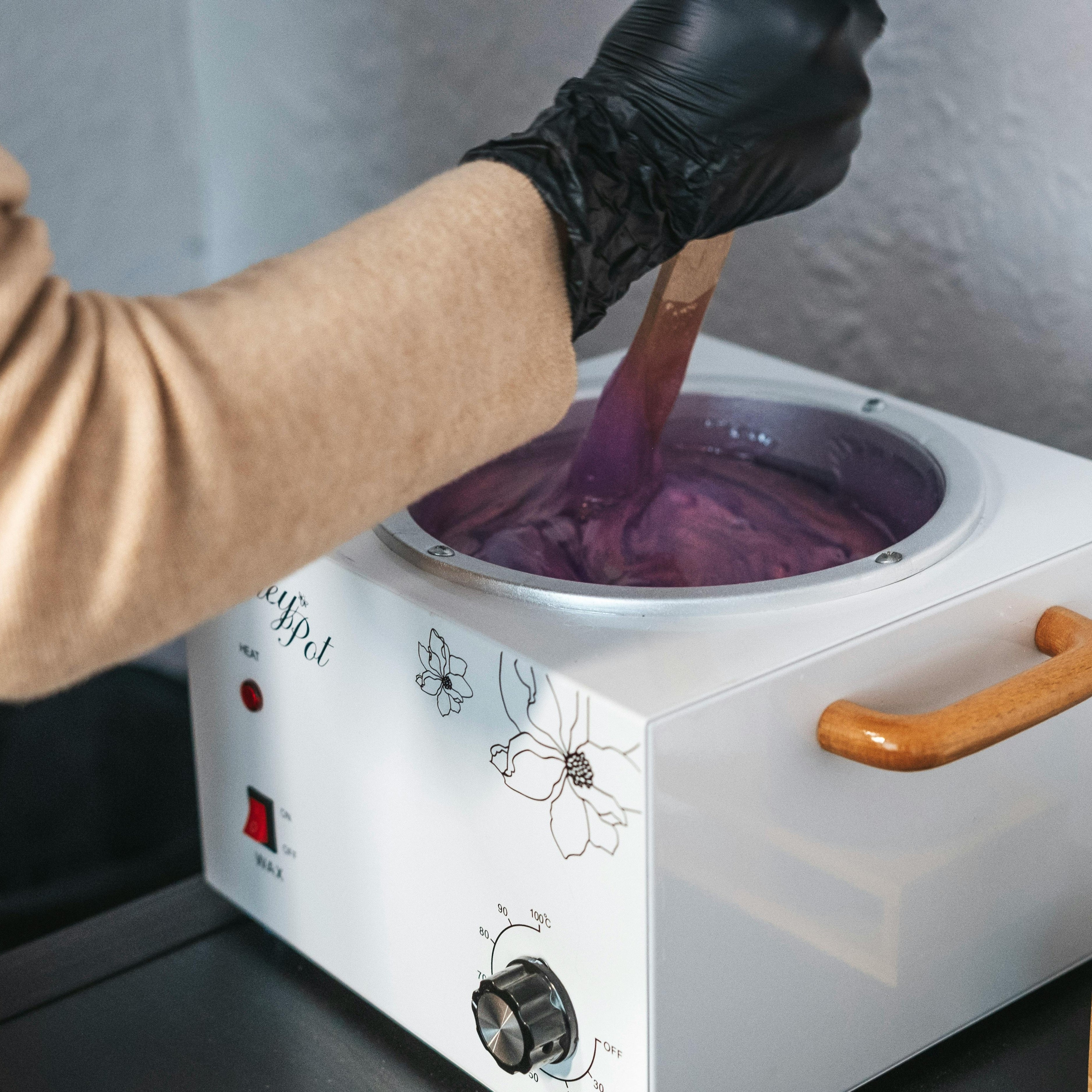 An esthetician wears a black glove and stirs a warming pot of hair removal wax