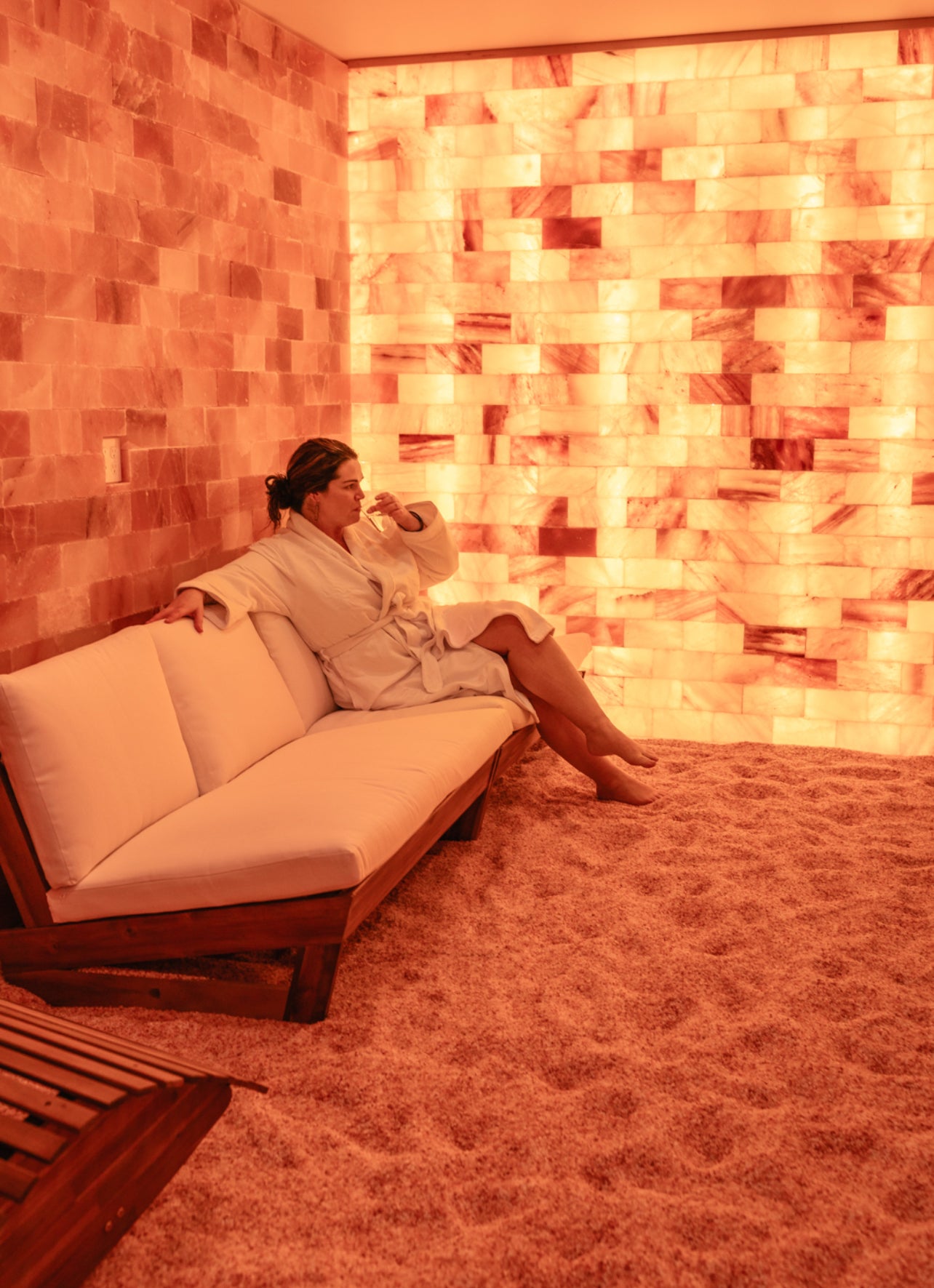 A woman sips water as she relaxes in a glowing, pink Himalayan Salt Vault in a luxury spa setting. 